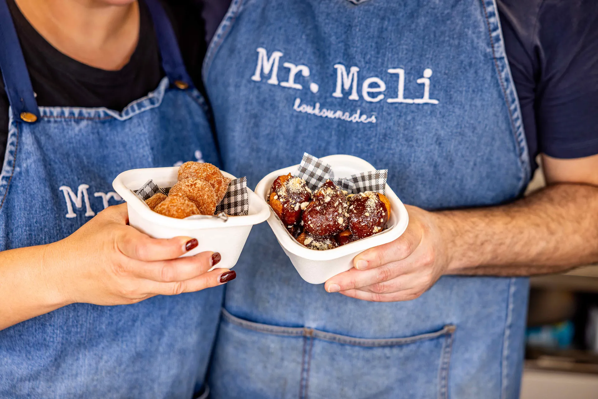 Two people wearing “Mr. Meli Loukoumades” aprons holding bowls of Greek doughnut desserts at the Perth Royal Show.