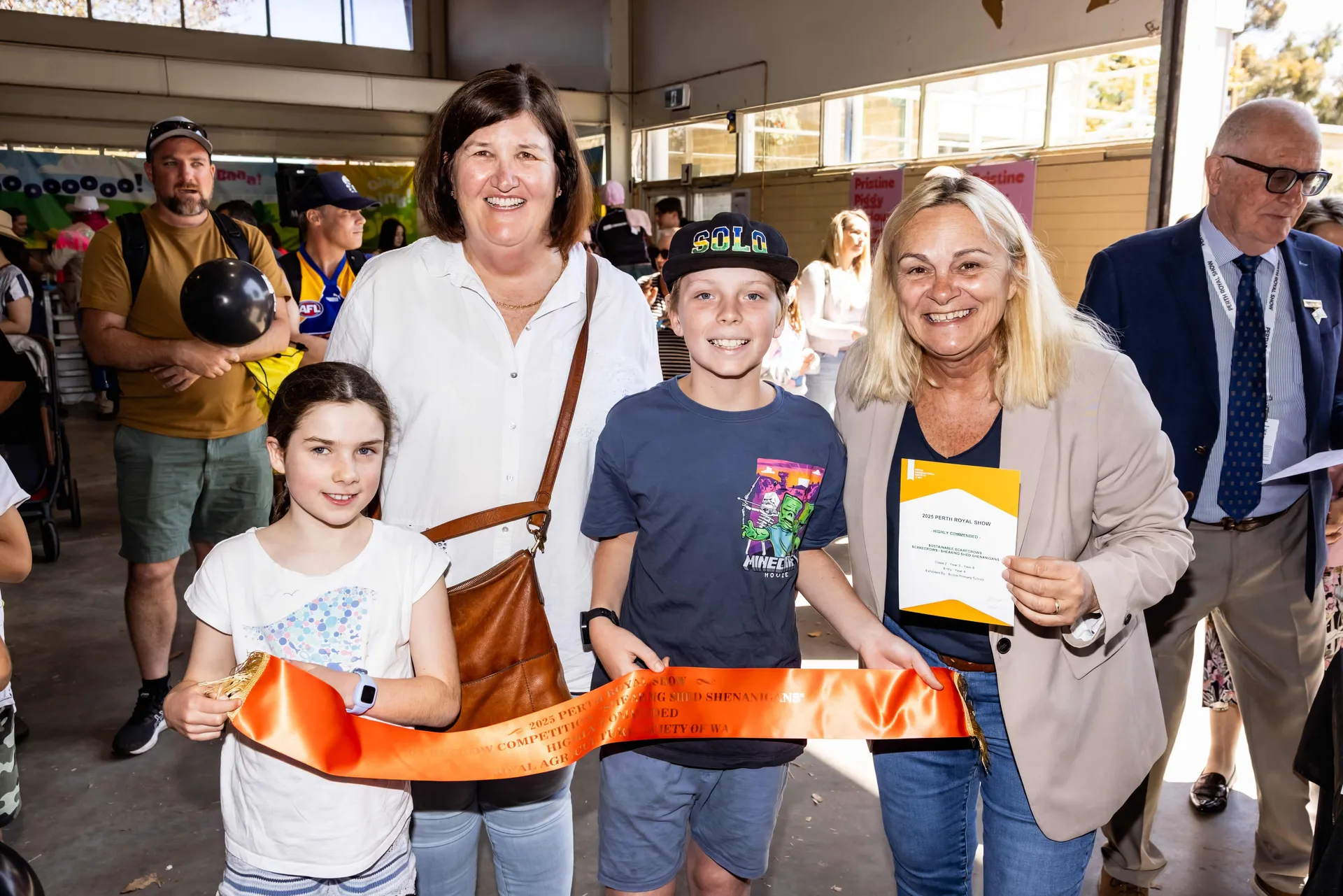 A group of smiling adults and children posing with an award ribbon and certificate inside a pavilion at the Perth Royal Show.