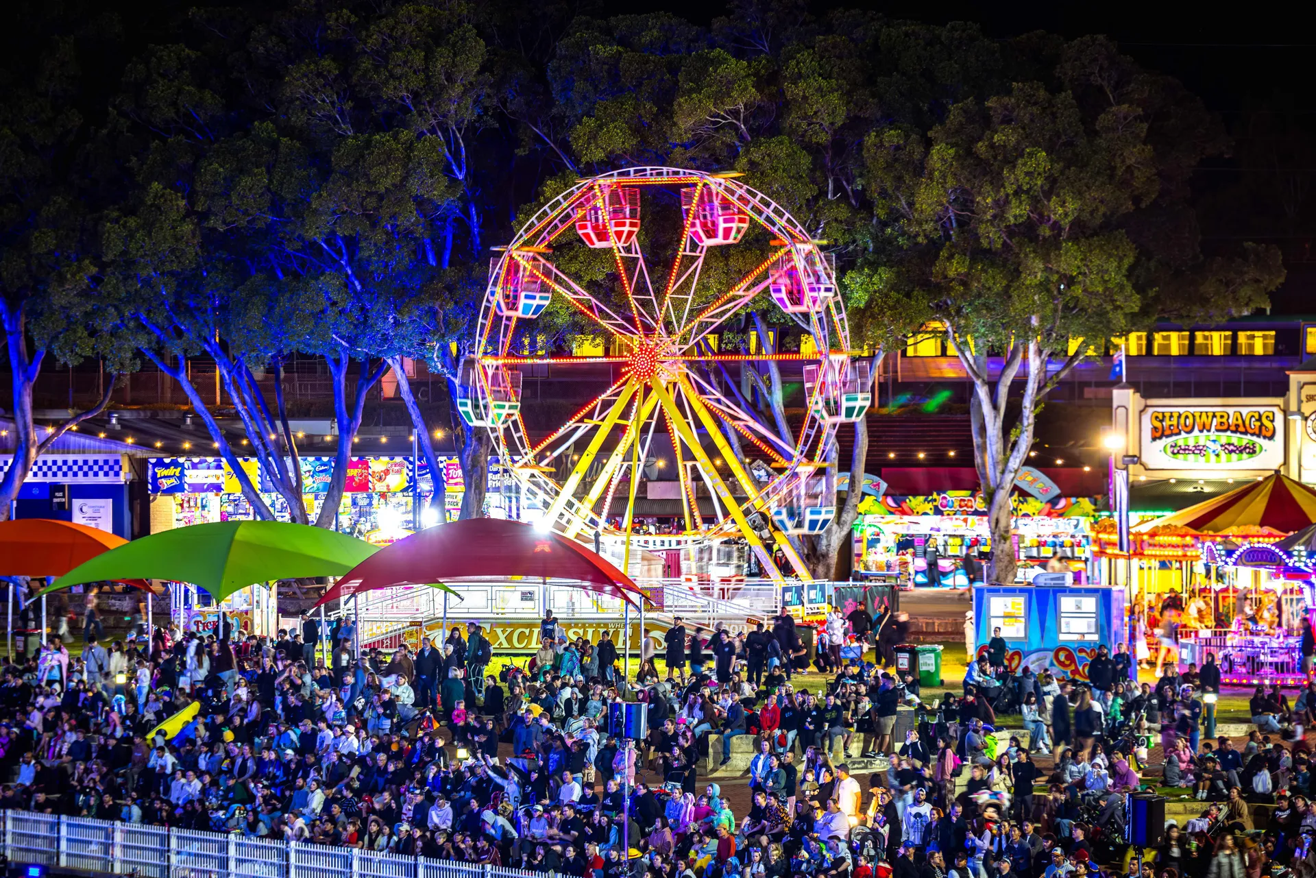 Crowds gathered at the Perth Royal Show at night, with a brightly lit ferris wheel and carnival rides in the background.