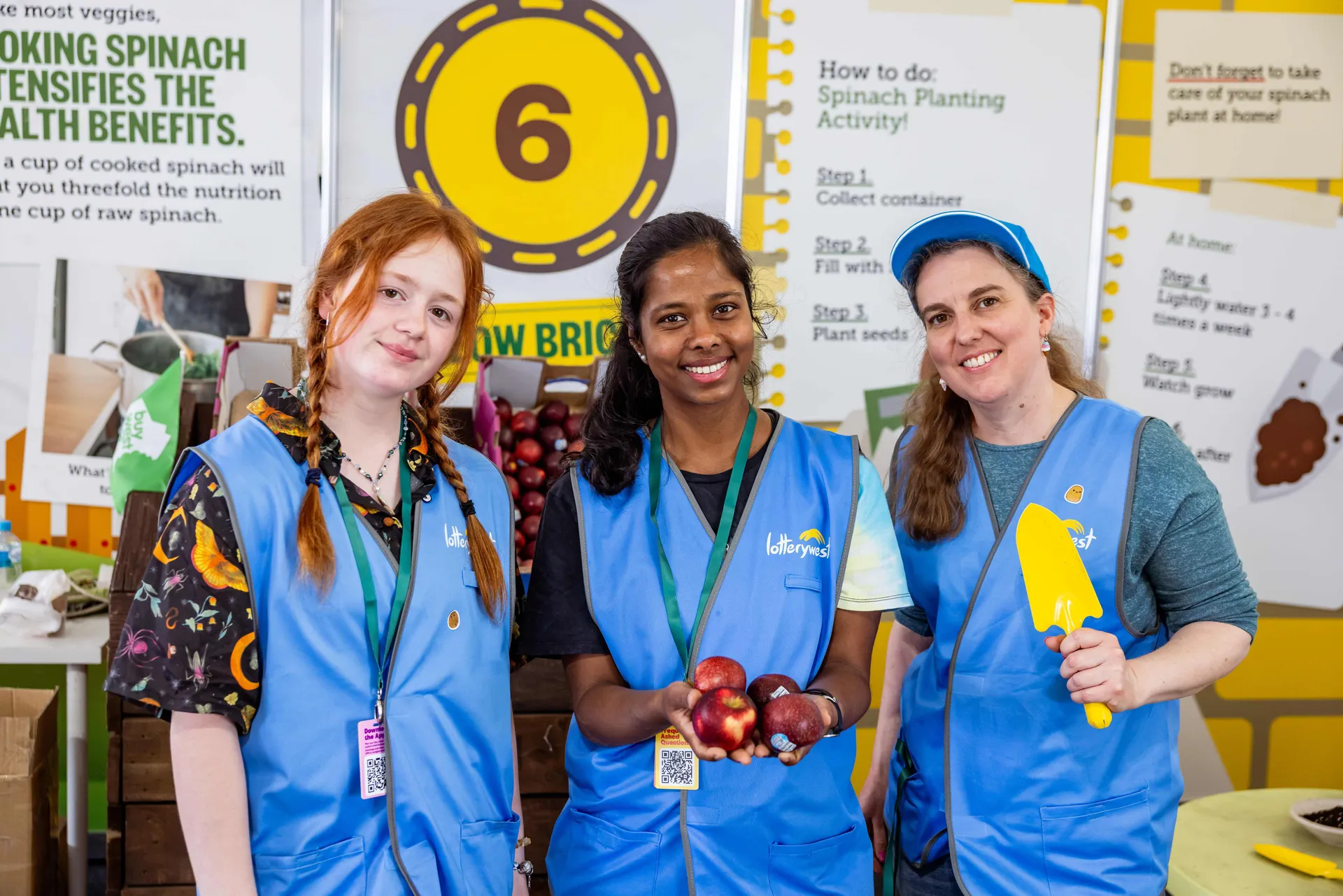 Volunteers at an educational activity booth at the Perth Royal Show holding apples and gardening tools.