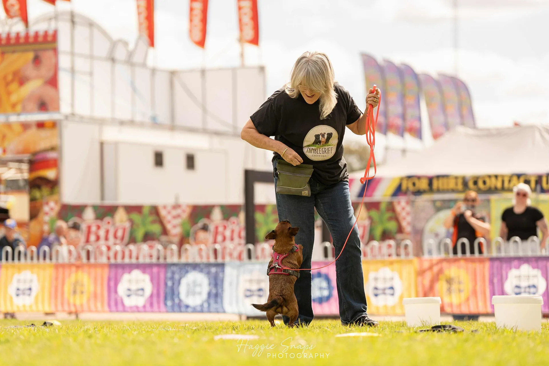 A woman training a small dog on a lead during an outdoor demonstration at the Perth Royal Show, with colourful stalls and spectators in the background.