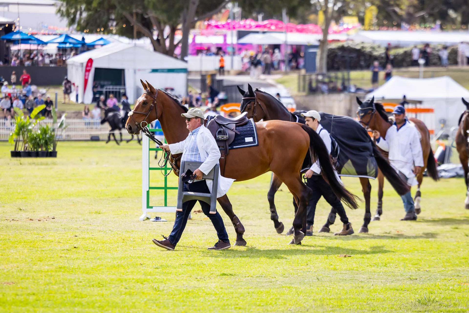 Handlers leading saddled horses across an arena during an equestrian event at the Perth Royal Show, with crowds and attractions visible in the background.