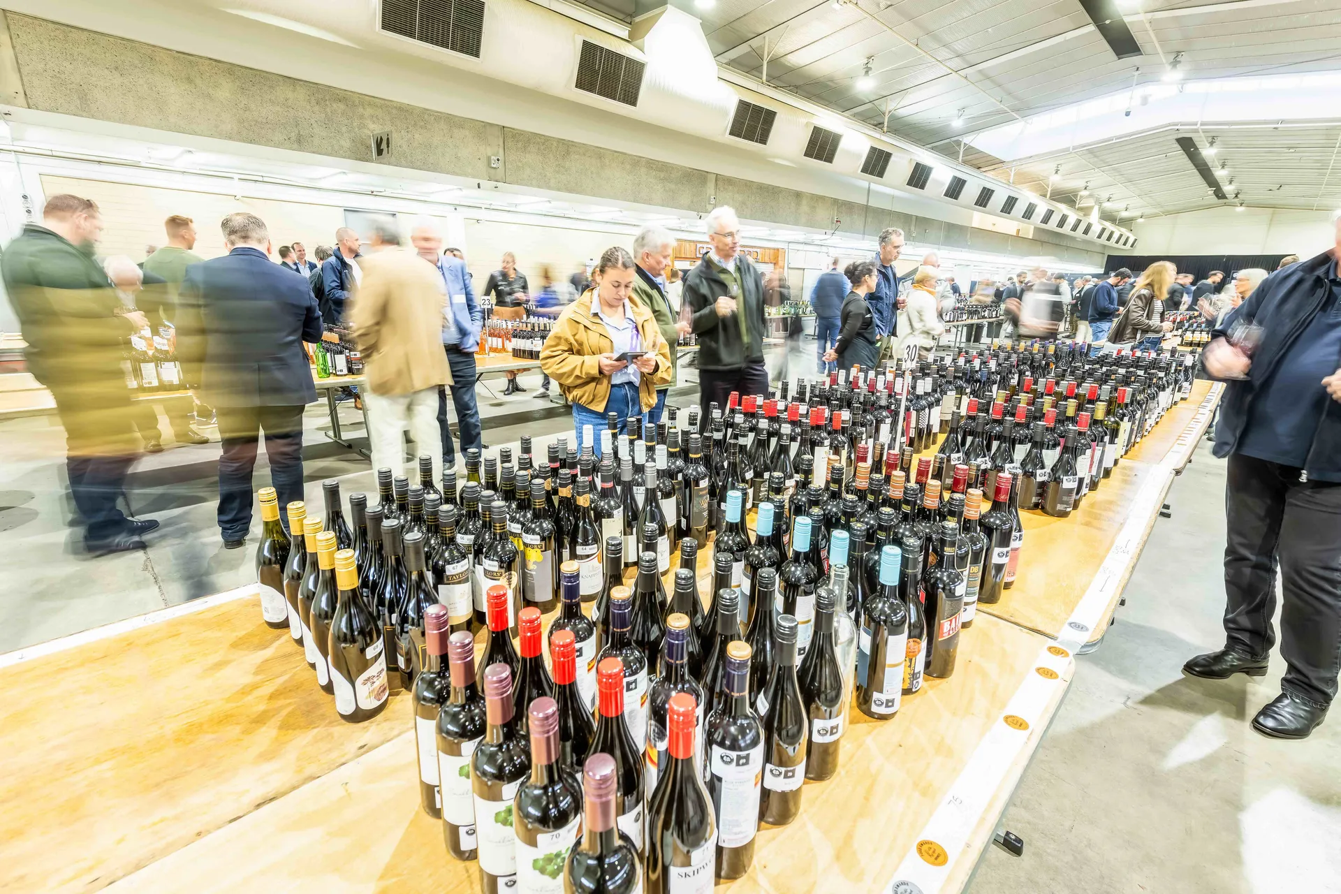 Rows of wine bottles lined up for judging at the Perth Royal Food Awards, with attendees tasting and reviewing entries in the background.