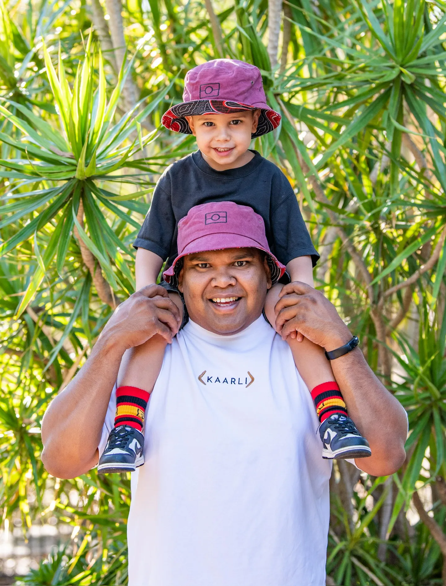 Man giving Piggy back to a little boy both wearing Aboriginal artists patterned bucket hats
