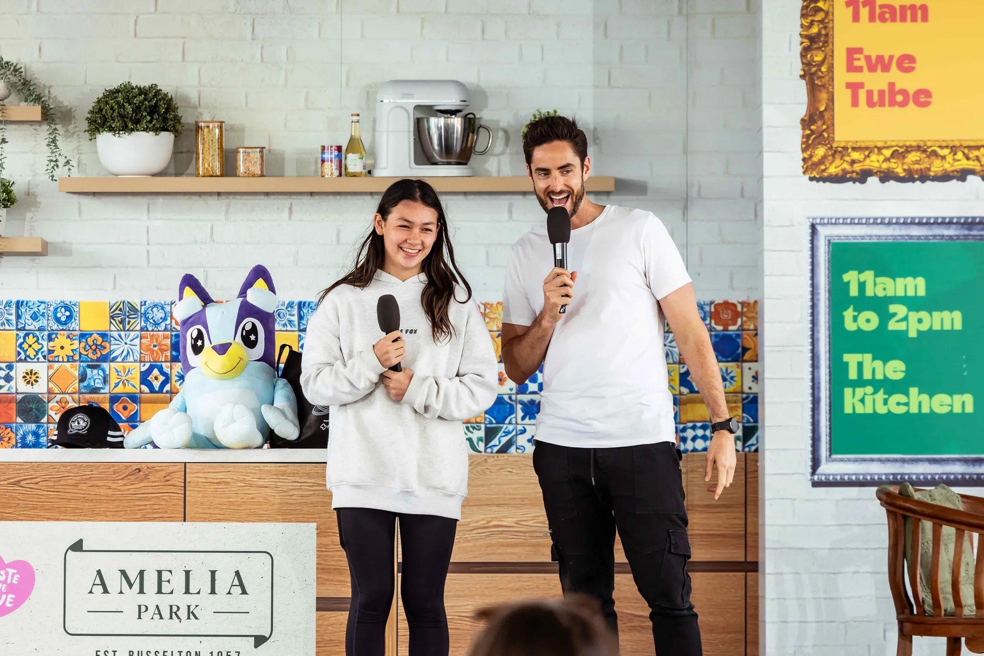 Two presenters holding microphones on stage at the Perth Royal Show, speaking to an audience in a kitchen-style setup with signage displaying event times in the background.