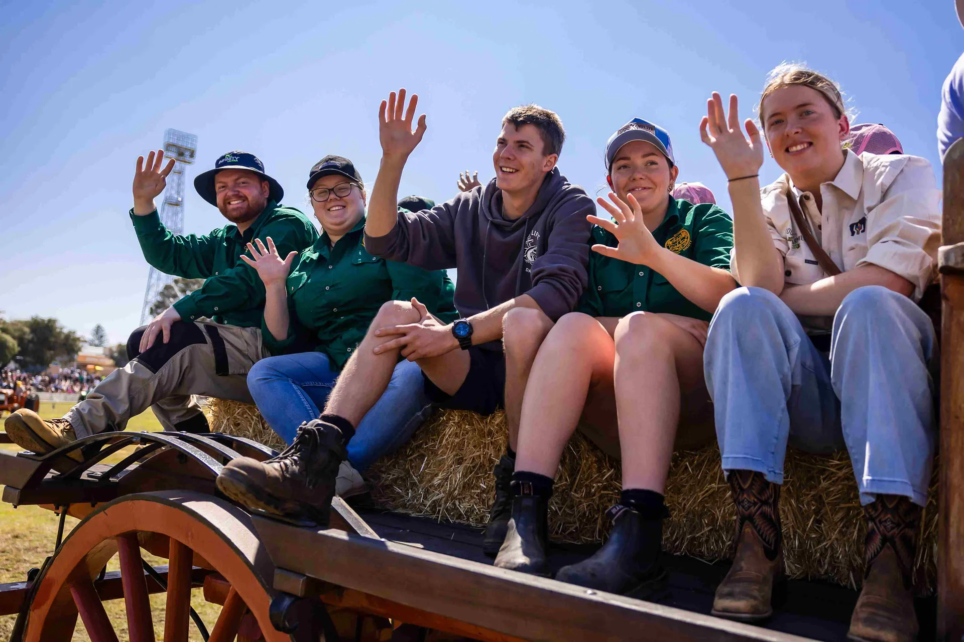 A group of smiling attendees wave from a hay bale wagon at Claremont Showground, enjoying the lively, community atmosphere.