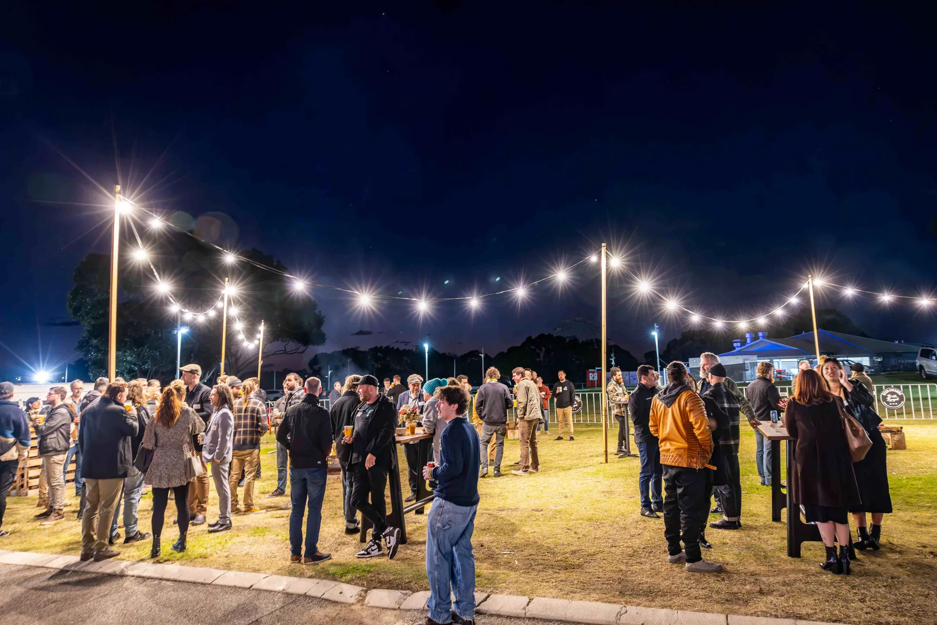 People socialising outdoors under string lights during an evening event at Claremont Showground.